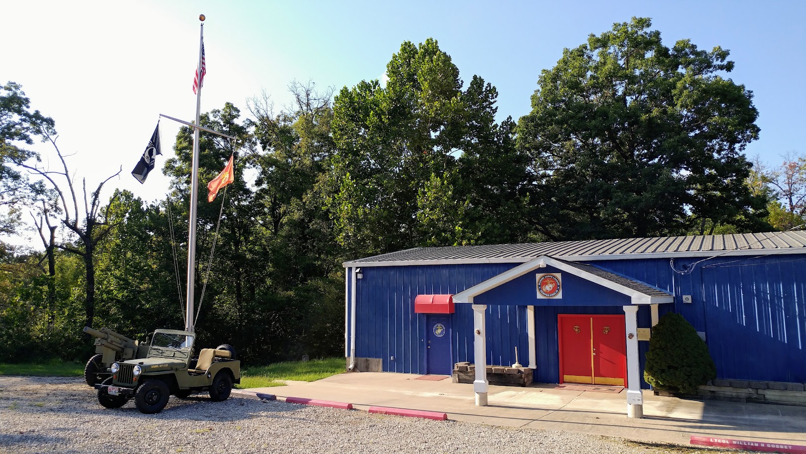 The front of the Marine Corps League Detachment 1137 building in Kaiser, Missouri - a blue metal building with red double doors under a white portico and the Marine Corps Eagle, Globe and Anchor emblem mounted above them. The American, POW/MIA and Marine Corps flags fly from a flagpole to the left, with a vintage military jeep and a towed howitzer parked beside it.